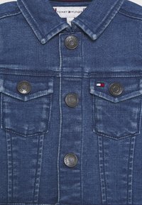 Kurtka dżinsowa w ciemnoniebieskim odcieniu, z zaostrzonym kołnierzem, dwoma przednimi kieszeniami oraz srebrzystymi guzikami z wytłoczonym logo.