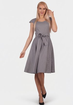 Blonde woman adjusting hair wearing a knee-length, short-sleeve, grey checkered dress with a waist tie and black heels, standing on white background.