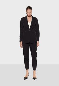 Black tailored blazer with a single button closure, embellished with gold-tone studs, paired with fitted black trousers and pointed-toe heels.