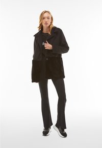 Abrigo negro con la mitad inferior de piel sintética texturizada, cierre de botones, cuello ancho y mangas largas, combinado con pantalones negros de campana y zapatillas.