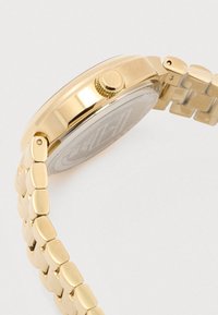 Złoty metalowy zegarek na rękę widoczny z boku, z teksturowaną koronką i ogniwowym paskiem na białym tle.