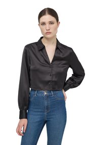 Blusa nera in satin cropped con scollatura a V, chiusura con bottoni e maniche lunghe a palloncino. Abbinata a jeans in denim blu a vita alta.