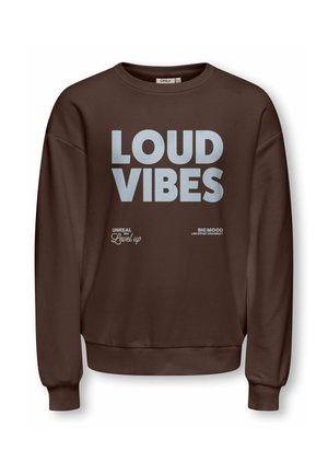 Usmievavá dievčina s kučeravými vlasmi stojí s rukou na bokoch, oblečená v hnedú mikinu s nápisom "LOUD VIBES,", sivé džínsy a biele topánky.