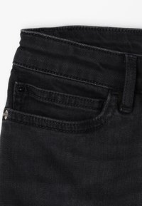 Los jeans de mezclilla negros presentan una textura suave, con un bolsillo delantero, lazos para el cinturón y un cierre de botón, mostrando un diseño clásico y ajustado.