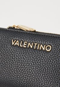 Portefeuille en cuir noir texturé avec le lettrage doré "VALENTINO" et une tirette de fermeture éclair dorée, affichant un design élégant et moderne.