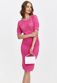 Różowa, dzianinowa sukienka z krótkimi rękawami, posiadająca teksturowany wzór, dopasowany krój oraz białą torebkę kopertową trzymaną w jednej ręce.