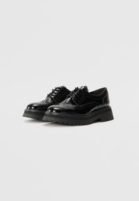 Black patent leather shoes with a wingtip design, featuring decorative brogue perforations, laces, and a chunky black rubber sole.