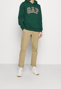 Felpa verde scuro con logo "GAP" in beige, tasca frontale, abbinata a pantaloni beige chiaro e sneakers bianche con accenti arancioni.