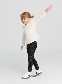 Top a maniche lunghe color crema con dettagli arricciati, abbinato a leggings neri con piccoli cuori dorati. Indossato con scarpe sportive bianche.