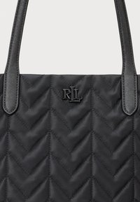 Crna protkana torba s uzorkom chevron, s crnim teksturiranim ručkama i metalnim logotipom. Glatka tekstura tkanine cijelom površinom.