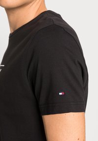 Hombre que lleva una camiseta negra de manga corta con un pequeño logo rectangular rojo, blanco y azul en la manga, sobre un fondo blanco.
