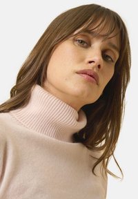 Vaaleanpunainen pooloneule, jossa on ribbikaulus, pehmeä rakenne ja rento istuvuus, kehystettynä luonnollisilla hiuksilla, jotka korostavat kasvoja.