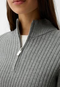 Szary prążkowany sweter z wysokim kołnierzem i srebrnym suwakiem. Materiał wydaje się miękki z widocznymi pionowymi paskami.