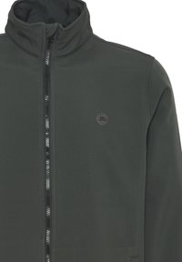 Dark green jacket with a high collar, textured fabric, full-length zipper, and a small circular logo on the chest.