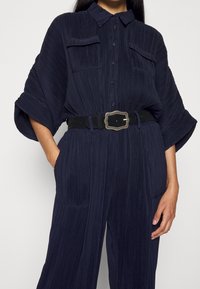 Femme portant une chemise plissée bleu marine à boutons avec deux poches poitrine, un pantalon plissé assorti et une ceinture noire avec une boucle décorative.
