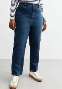 Jeans blu scuri a vita alta con una vestibilità dritta, dotati di chiusura con bottone anteriore e tasche frontali, abbinati a sneaker bianche.