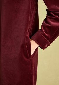 Burgunderrote Samtbluse mit glatter Textur, ausgestattet mit einer Seitentasche und Knopfmanschetten. Nahaufnahme des Ärmels.