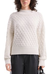 Cremefarbener gestrickter Pullover mit einem strukturierten Rautenmuster, geripptem Saum und Bündchen sowie einem hohen, abgerundeten Kragen. Kombiniert mit schwarzer Hose.