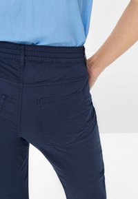 Marineblauwe elastische broek met twee achterzakken, voorzien van vlakke naden en een gladde textuur, gedragen met een lichtblauw shirt.