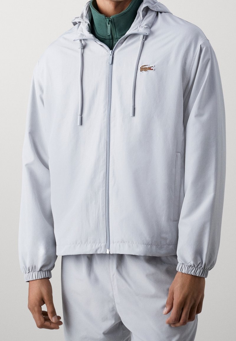 Chaqueta gris claro con cremallera y capucha, puños elásticos y logo de Lacoste. Hecha de una tela suave, presenta un ajuste relajado y bolsillos laterales.