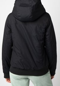 Personne portant une veste noire à capuche unie vue de dos, avec un pantalon vert clair sur un fond neutre.