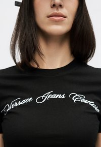 Γυναίκα που φοράει μαύρο μπλουζάκι με λευκά καλλιγραφικά γράμματα "Versace Jeans Couture" στο στήθος, με σκούρα ίσια μαλλιά μήκους έως τους ώμους ορατά.