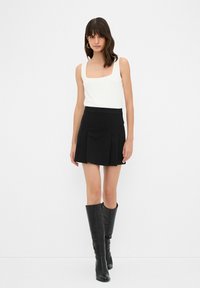 Un débardeur blanc à encolure carrée associé à une mini-jupe plissée noire. Le mannequin porte des bottes en cuir noires montantes. Design simple et moderne.