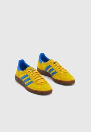 Paire de baskets jaunes en daim avec des bandes bleues, une doublure intérieure bleue, des lacets jaunes et des semelles en caoutchouc marron sur fond gris.