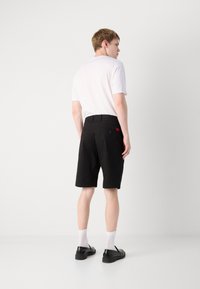 Shorts negri cu o textură netedă, având un buzunar la spate și o etichetă roșie cu logo. Modelul poartă o cămașă albă și pantofi negri cu șosete albe.
