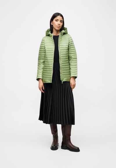 Femme portant une doudoune vert clair, une jupe midi plissée noire, un haut noir et des bottes marron montant jusqu'aux genoux, debout devant un fond blanc uni.