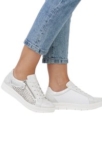 Baskets blanches avec un mélange de matériaux lisses et texturés, dotées d'un design à découpes décoratives et d'un zip sur le côté. Semelles blanches épaisses.