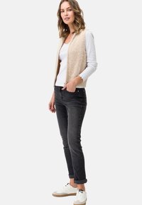Gilet in finta pelliccia beige con zip frontale, indossato sopra una camicia bianca; abbinato a jeans scuri e sneakers bianche con accenti beige.
