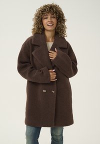 Pruun topeltkäänatud mantel, valmistatud tekstuurilisest kangast, millel on silmapaistvad kraed ja suurendatud varrukad. Stiliseeritud kerge pluusi ja teksade peale.