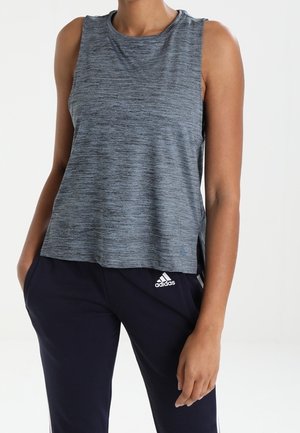 Frau trägt ein ärmelloses graues Sportoberteil und marineblaue Adidas-Trainingshose, steht mit einer Hand in der Tasche vor weißem Hintergrund.