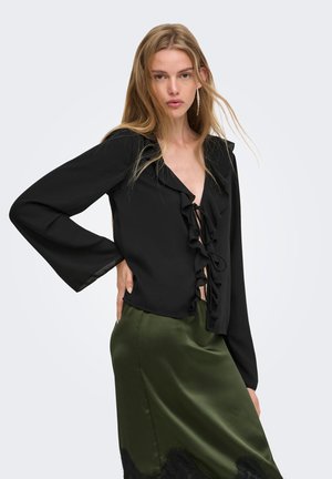 Femme aux longs cheveux blonds porte un chemisier noir à volants avec un lien devant et une jupe en satin vert olive, posant avec la main sur la hanche, fond neutre.