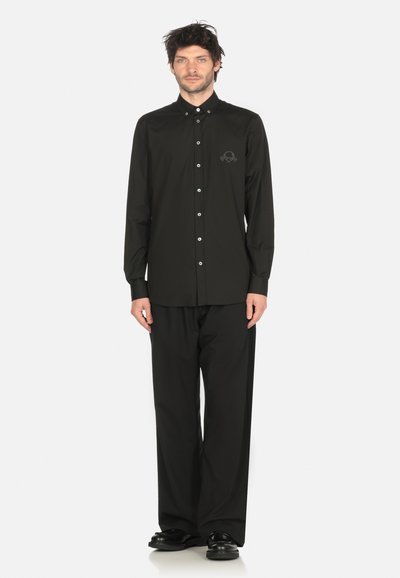 Uomo in piedi con un outfit completamente nero, composto da una camicia a maniche lunghe con bottoni e un piccolo logo ricamato, pantaloni larghi e scarpe nere.