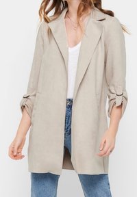 Kvinna som bär en ljusbeige kavaj med öppna framkanter och uppvikta ärmar över en vit topp och blå jeans, som visar midjan och en del av nacken.