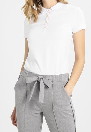 Femme portant un polo blanc à manches courtes et un pantalon gris taille haute avec une ceinture en tissu nouée et des poches.