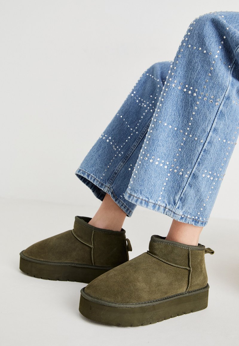 Stivaletti alla caviglia in suede verde oliva con suola spessa e testurizzata e linguetta. Abbinati a jeans in denim azzurro chiaro con dettagli decorativi.