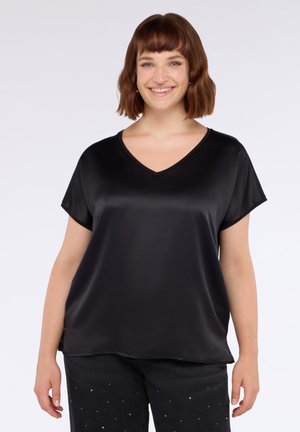 Lachende vrouw met kort bruin haar, gekleed in een zwarte satijnen V-hals korte mouw blouse en zwarte broek met kleine witte stippen.