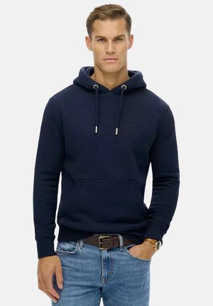 Mann trägt navyblauen Hoodie und hellblaue Jeans mit braunem Gürtel und Armbanduhr, steht vor einfarbigem Hintergrund.