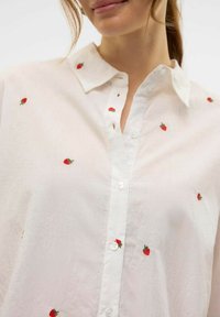 Camicia bianca con bottoni, decorata con piccoli ricami di fragole rosse, dotata di un colletto classico e realizzata in un tessuto leggero e leggermente strutturato.
