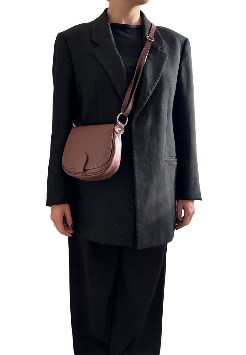 Borsa a tracolla in pelle marrone con forma arrotondata, pattina pieghevole e tracolla regolabile, indossata sopra un blazer nero oversize e pantaloni.