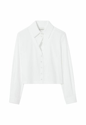 Femme portant un chemisier blanc boutonné, une jupe midi noire avec des volants, une ceinture chaîne dorée, des talons noirs à brides, tenant un petit sac marron, debout sur fond gris.