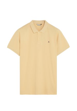 Polo de manga corta beige con dos botones y un pequeño logotipo oscuro de calavera y huesos cruzados en el lado izquierdo del pecho.