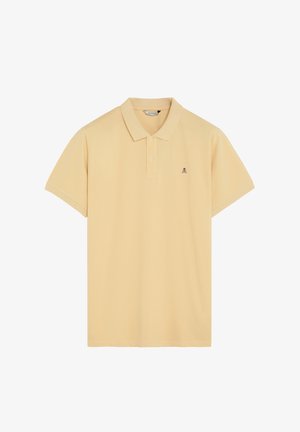 Polo de manga corta beige con dos botones y un pequeño logotipo oscuro de calavera y huesos cruzados en el lado izquierdo del pecho.