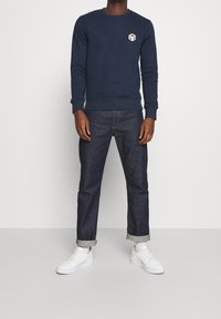 Man som bär en marinblå sweatshirt med en liten geometrisk logotyp, mörka upprullade jeans, vita strumpor och vita sneakers som står mot en enfärgad bakgrund.