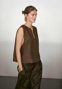 Brown linen sleeveless top with a round neckline and a front zip detail, paired with brown loose-fitting trousers and a metallic bracelet.