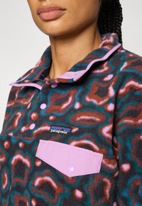 Fleece-genser med et brunt, rosa og teal abstrakt mønster. Har skjorte-krage og en rosa lommepose med trykknapp. Patagonia-logoen er synlig.