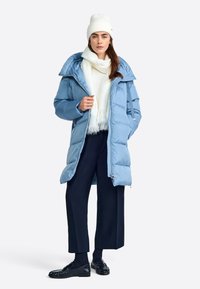 Manteau rembourré bleu clair avec une capuche, doté d'une fermeture éclair à l'avant, de poches latérales et de détails matelassés. Associé à une écharpe blanche et à un pantalon sombre.